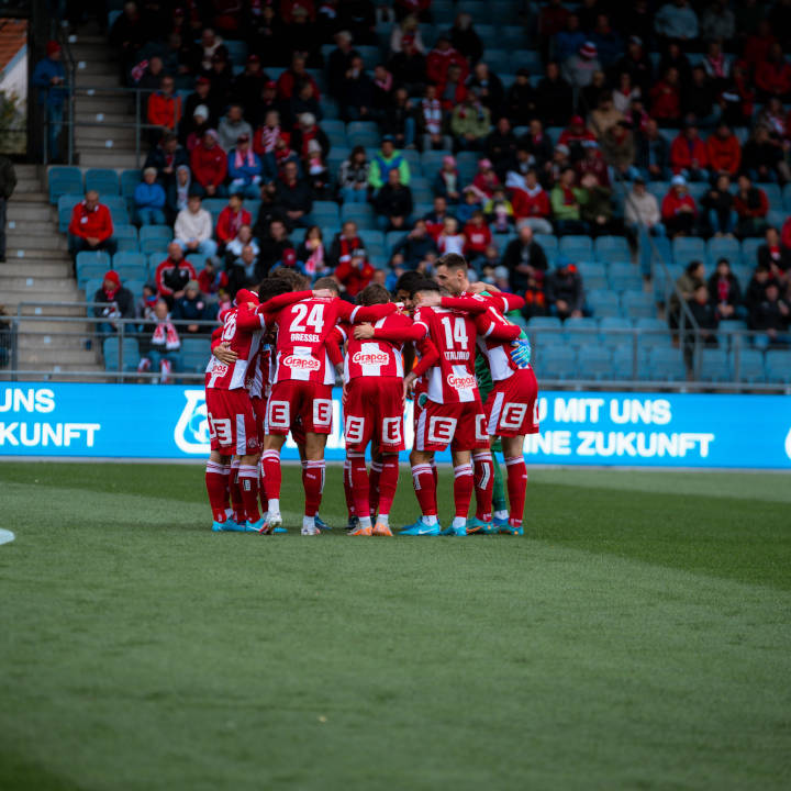 Eine Fussballmannschaft bespricht sich vorm Spiel. Sie stehen im Kreis und haben sich die Arme gegenseitig über die Schultern gelegt.